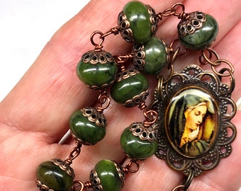 Könnte beinhalten: Ein grünes Steinrosenkranz-Armband mit einer Kupferkette und einem Kupferanhänger mit einem religiösen Bild. Der Anhänger hat ein filigranes Design um das Bild herum.