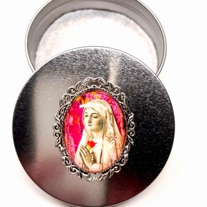 May include: A silver metal box with a round lid. The lid features a silver-toned oval-shaped cameo with a colourful image of the Virgin Mary holding a red rose.