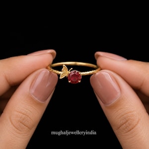 May include: A gold ring featuring a round, faceted red gemstone set next to a small, detailed gold butterfly motif. The ring band has a textured, hammered finish. The text mughaljewelleryindia is printed at the bottom of the image.