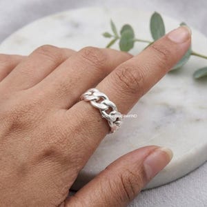 Puede incluir: Un anillo de plata con un diseño de eslabones de cadena tipo barbada pulidos. La banda consta de eslabones metálicos redondeados e interconectados que envuelven el dedo, creando un patrón continuo y texturizado con un acabado metálico brillante.