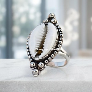 Peut inclure: Une bague en argent avec un coquillage cauri blanc central serti dans un cadre en métal décoratif avec une bordure perlée. La bague possède un anneau en argent poli et lisse, avec le texte ShreeJaipurSilverIND imprimé sur la coquille.