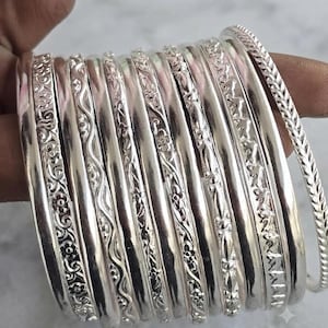 Op de afbeelding: Een set van negen zilverkleurige armbanden met diverse reliëfpatronen, waaronder bloemmotieven, golvende lijnen en geometrische texturen. Elke armband heeft een gepolijste metalen afwerking en één armband heeft een gevlochten touwontwerp.