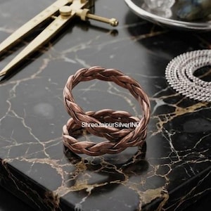 Pode incluir: Um anel em tom de cobre com um design trançado e uma superfície de metal texturizada e torcida. O anel está posicionado centralmente, com o texto ShreeJaipurSilverIND visível na superfície. O metal tem um acabamento quente castanho-avermelhado.