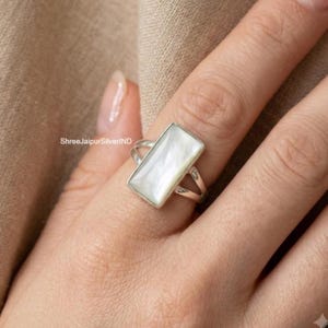 Può includere: Un anello in argento con un inserto rettangolare in madreperla iridescente. La fascia ha un design diviso che si collega alla montatura che sostiene la pietra. Il testo ShreeJaipurSilverIND è visibile sul lato della montatura dell'anello.