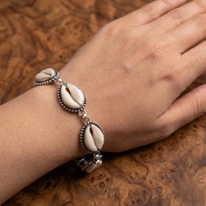 Könnte beinhalten: Ein Armband mit einer Reihe weißer Kaurimuscheln, die jeweils in eine dekorative, silberfarbene Metallfassung mit Perlenrand eingefasst sind. Die Muscheln sind durch kleine silberfarbene Metallringe zu einer Kette verbunden.