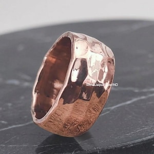 Può includere: Un anello largo in rame lucido caratterizzato da una distinta texture martellata su tutta la superficie esterna. Il metallo presenta una calda tonalità bruno-rossastra metallica con riflessi luminosi. Il testo ShreeJaipurSilverIND è visibile.