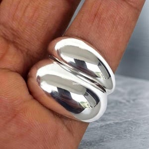 May include: A polished silver ring featuring two smooth, rounded, bulbous bands that are joined together. The surface of the metal is highly reflective, showing clear highlights and shadows. The text ShreeSilverIND is visible on the ring.