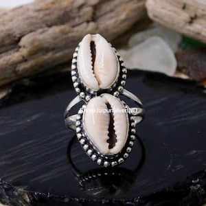 May include: A silver ring featuring two vertically stacked white cowrie shells. Each shell is encased in a beaded silver setting. The band of the ring is split into two thin silver wires that connect to the central shell design. The text ShreeJaipurSilverIND is visible.