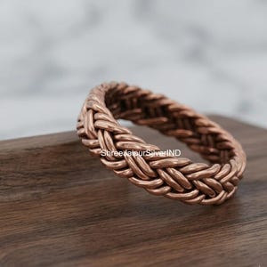 Op de afbeelding: Een koperen ring met een doorlopend gevlochten of geweven metaalpatroon over de gehele band. Het oppervlak van de ring vertoont een metallic roodbruine afwerking met complexe in elkaar grijpende strengen die een getextureerd uiterlijk creëren.
