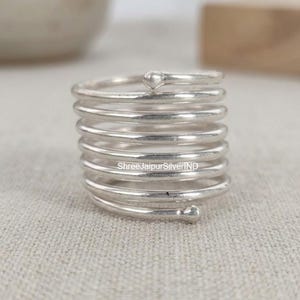 May include: A silver-colored metal ring featuring a multi-coil spiral design with a rounded bead at each end. The text ShreeJaipurSilverIND is visible on the surface of the metal band. The ring is constructed from a single continuous wire.