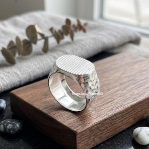 Peut inclure: Une bague en argent avec un dessus plat de forme ovale orné d'un motif à rayures linéaires texturées. L'anneau de la bague présente une finition martelée et alvéolée, créant une surface réfléchissante. La bague est faite d'une seule pièce.