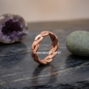 Può includere: Un anello in rame caratterizzato da un design in metallo intrecciato e ritorto. La fascia presenta una calda finitura metallica color bruno-rossastro con un aspetto intrecciato e testurizzato. Il testo ShreeJaipurSilverIND è visibile.