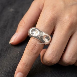 Può includere: Un anello in argento lucido con una fascia spessa e arrotondata caratterizzata da due distinte rientranze concave e lisce sulla parte anteriore. Il metallo ha una finitura lucida e riflettente che cattura la luce, creando un aspetto liscio e solido.