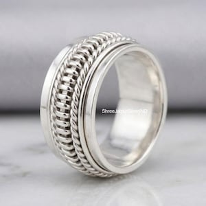 May include: A wide silver spinner ring featuring a central band with a twisted rope texture flanked by two smooth, polished silver edges. The ring displays the text ShreeJaipurSilverIND on its inner surface, showcasing a metallic finish.