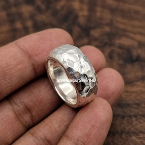 May include: A wide silver ring featuring a textured, hammered surface with a polished finish. The band has a rounded profile and a reflective metallic sheen. The text ShreeJaipurSilverIND is visible on the inner surface of the ring band.