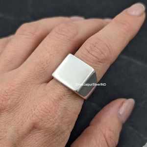 May include: A polished silver signet ring featuring a flat, square-shaped face and a smooth, solid band. The metal surface is reflective and plain, with no engravings or stones. The text ShreeJaipurSilverIND is visible on the side of the ring.