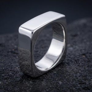 Könnte beinhalten: Ein polierter silberfarbener Ring mit einem markanten quadratischen Band, abgerundeten Ecken und einer glatten, reflektierenden Metalloberfläche. Der Ring hat ein breites, flaches Oberprofil und eine hohle Mitte, was eine klare, solide Bauweise zeigt.