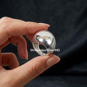 Può includere: Un anello in argento lucido con una superficie superiore liscia, arrotondata e a forma di cupola. L'interno della fascia presenta il testo inciso 925 D-85. L'anello ha una finitura metallica molto brillante e una struttura spessa e solida.