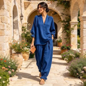 May include: A deep blue two-piece set consisting of a long-sleeved tunic with a V-neckline and ruffled cuffs, paired with matching loose-fitting trousers featuring gathered, ruffled hems. The fabric has a textured appearance.