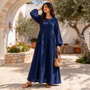 May include: A long-sleeved navy blue maxi dress with a round neckline and a gathered waistline. The dress features a loose, flowing skirt that reaches the ankles. A small, round, woven straw handbag with brown leather handles is held in one hand.