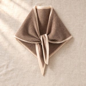 May include: A triangular knit scarf in a solid medium brown color featuring a contrasting cream-colored border along all edges. The scarf is folded into a point with a central knot detail, showcasing a soft, textured fabric construction throughout.