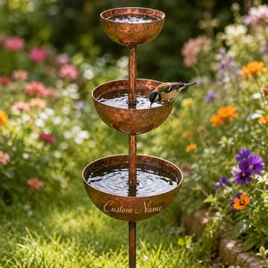 May include: A three-tiered copper bird bath featuring a hammered metal finish and a central vertical support pole. The lowest basin is engraved with the text Custom Name. A small bird is perched on the edge of the middle basin drinking water.