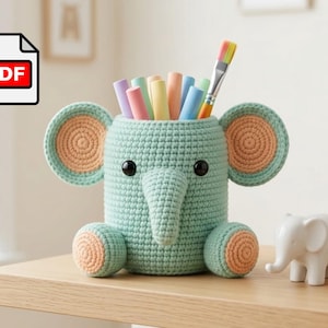 Op de afbeelding: Een mintgroene gehaakte olifant-pennenhouder met grote perzikkleurige binnenoren en voetkussentjes, zwarte plastic ogen en een slurf. De houder bevat diverse pastelkleurige krijtjes en een penseel. Een klein wit olifantfiguurtje staat ernaast.