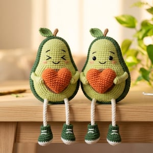 May include: Two crocheted avocado plush toys with light green centres and dark green outer skins. Each avocado has a smiling face, a small brown stem, and holds a textured orange heart. They have long dangling legs with dark green shoes.