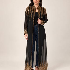 May include: A long, black, sheer duster jacket featuring a mandarin collar and intricate gold sequin embellishments that radiate from the shoulders and hemline. The open-front garment has long sleeves with matching gold sequin detailing at the cuffs.