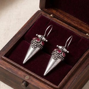 Puede incluir: Un par de pendientes colgantes de tono plateado con forma cónica y base puntiaguda. Los pendientes están decorados con pequeñas piedras rojas, intrincados detalles de cuentas metálicas y un motivo floral. Se presentan dentro de una caja de madera oscura.