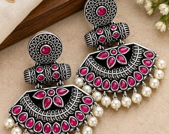 May include: A pair of oxidized silver-tone earrings featuring a circular top stud and a large semi-circular pendant. The design is accented with vibrant pink teardrop-shaped stones and small white faux pearls dangling from the bottom edge.