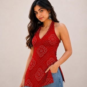May include: A sleeveless red halter top featuring a white dotted pattern with repeating floral-like motifs and side slits, paired with light blue denim jeans. The top has a deep V-neckline and a fitted silhouette that extends to the hips.