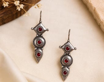 May include: A pair of drop earrings crafted from oxidized silver-tone metal, each featuring a diamond-shaped top, a circular middle section, and a pointed bottom, all accented with small, round, deep red stones set in the center of each shape.
