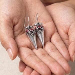 May include: A pair of silver-toned drop earrings featuring a conical shape with an oxidized finish. Each earring is adorned with small, round, deep red stones set in a decorative band above a floral-patterned metal detail and a hook wire.
