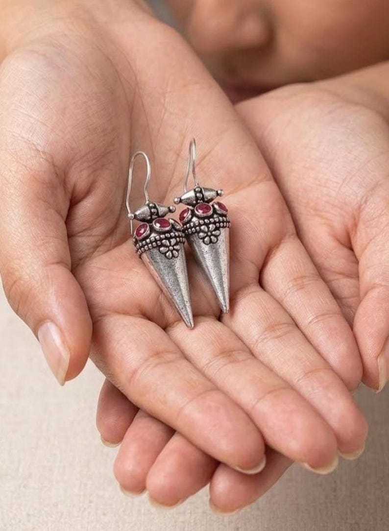 May include: A pair of silver-toned drop earrings featuring a conical shape with an oxidized finish. Each earring is adorned with small, round, deep red stones set in a decorative band above a floral-patterned metal detail and a hook wire.