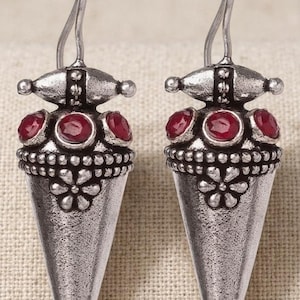 May include: A pair of silver-toned drop earrings featuring a conical shape with an oxidized finish. Each earring is adorned with three small, round, deep red stones set near the top and a raised floral pattern embossed on the lower section.