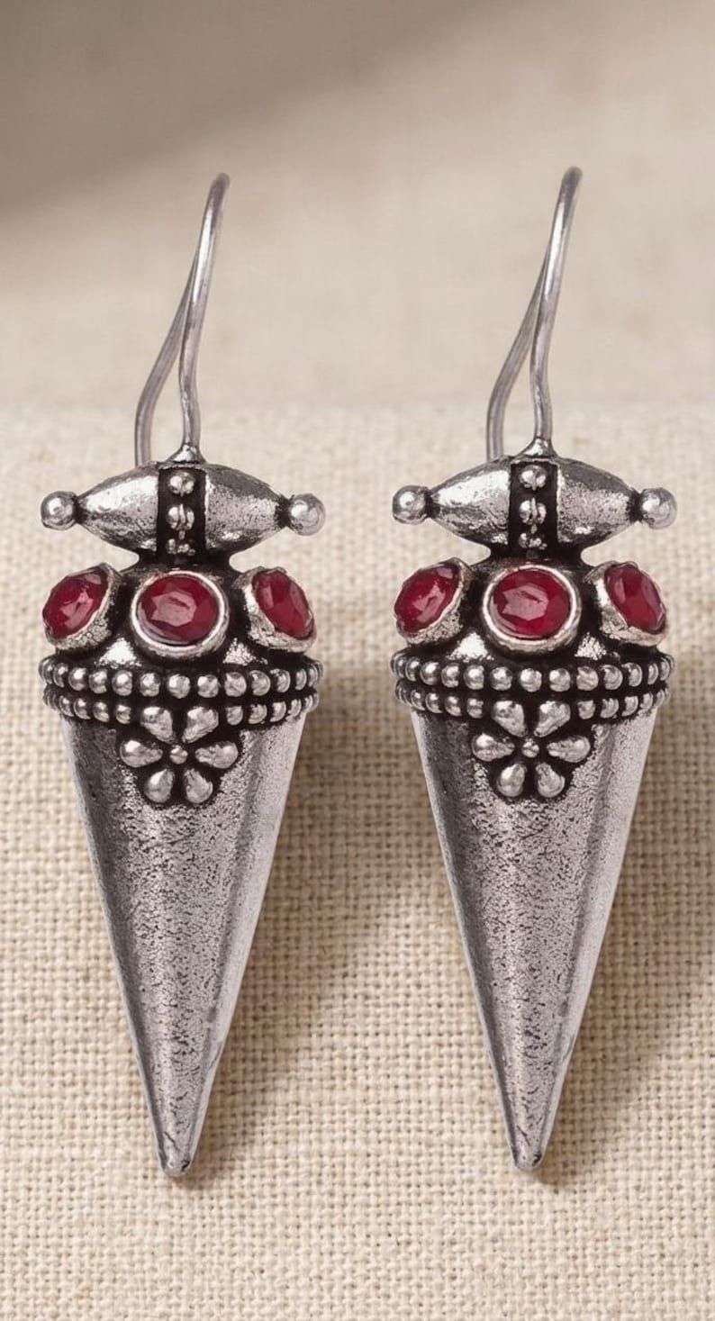 May include: A pair of silver-toned drop earrings featuring a conical shape with an oxidized finish. Each earring is adorned with three small, round, deep red stones set near the top and a raised floral pattern embossed on the lower section.
