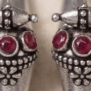 May include: A pair of silver-toned drop earrings featuring a conical shape with a textured, oxidized finish. Each earring is adorned with two round, faceted deep red stones and a small embossed floral pattern near the base, suspended by a hook.