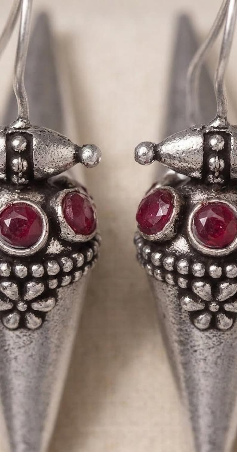May include: A pair of silver-toned drop earrings featuring a conical shape with a textured, oxidized finish. Each earring is adorned with two round, faceted deep red stones and a small embossed floral pattern near the base, suspended by a hook.
