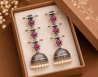 May include: A pair of silver-toned dangle earrings featuring three vertical magenta stones set in metal frames, ending in a textured bell-shaped jhumka base adorned with a row of small white beads, presented inside a brown cardboard box.