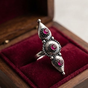 Peut inclure: Une bague argentée au design vertical allongé, ornée de trois pierres précieuses circulaires rose foncé serties dans du métal texturé. La pierre centrale est entourée d'une bordure perlée, tandis que les pierres aux extrémités sont pointues.