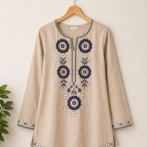 Könnte beinhalten: Eine beige langärmelige Tunika mit geschlitztem Ausschnitt und dunkelblauer sowie weißer Blumenstickerei auf der Vorderseite. Das Kleidungsstück hat passende Stickereien an den Ärmelbündchen und am abgerundeten Saum sowie ein Etikett mit der Aufschrift KAYA.