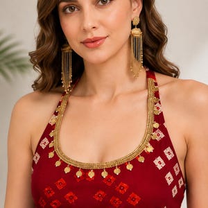 May include: A maroon halter-neck top featuring a white geometric pattern and a gold-toned trim adorned with small hanging circular discs. The garment is paired with long, gold-toned jhumka-style earrings consisting of a stud and multiple chains.