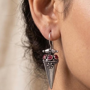 May include: A silver-toned drop earring featuring a conical pendant with a textured floral motif at the top. The design includes three small, round, deep red stones set horizontally above the cone, suspended from a simple curved silver wire hook.