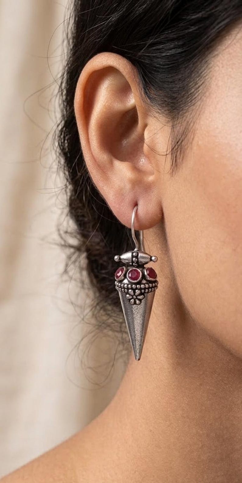 May include: A silver-toned drop earring featuring a conical pendant with a textured floral motif at the top. The design includes three small, round, deep red stones set horizontally above the cone, suspended from a simple curved silver wire hook.