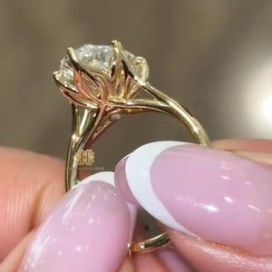 May include: A gold ring featuring a large, round, brilliant-cut center stone held by a six-prong setting. The prongs are shaped like petals, extending from a polished gold band. A small logo with the letters HH is visible on the side of the setting.