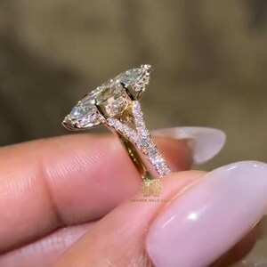 Puede incluir: Un anillo de oro con una gran piedra central de talla marquesa rodeada por un halo de piedras más pequeñas. La banda está dividida en forma de V y está incrustada con piedras en pavé. El texto HEAVEN HALO CO es visible en el engaste.