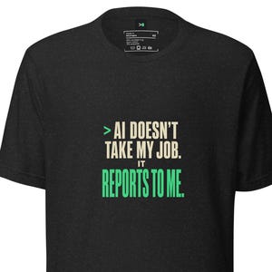 Puede incluir: Una camiseta de manga corta gris carbón con el texto en blanco y verde: >AI DOESN'T TAKE MY JOB. IT REPORTS TO ME. La camiseta tiene un cuello redondo y un corte estándar, con una pequeña etiqueta de marca visible en el interior del cuello.