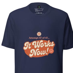 Op de afbeelding: Een marineblauw t-shirt met korte mouwen en een grafische print met een kleine bloem, de tekst "Attempt 47 of 47... It Works Now!" in wit en oranje, en een klein oranje smiley-icoontje in het midden op de borst van het kledingstuk.