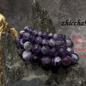 May include: A collection of round, polished purple amethyst beads arranged in four distinct strands. The text zhiccha99 is visible, along with labels indicating bead sizes of 6mm, 8mm, 10mm, and 12mm. The beads feature natural color variations.